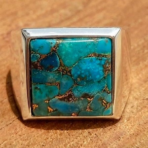 May include: A square silver ring featuring a large, turquoise-colored stone with a mosaic pattern of blue and brown hues. The stone is set within a polished silver band, creating a bold and eye-catching design.