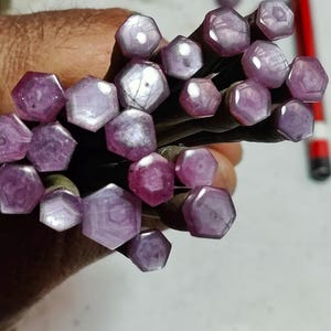 May include: A collection of hexagonal-shaped, polished gemstones in varying shades of purple and pink. The gemstones are held together, with a pencil visible in the background. The gemstones have a translucent quality, with visible internal patterns.
