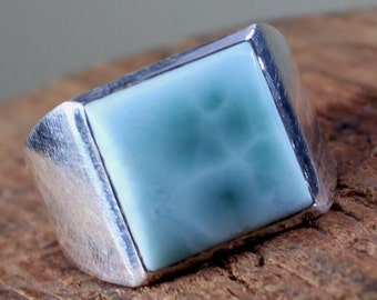 Può includere: Un anello d'argento con una pietra quadrata di colore blu chiaro. La pietra ha un motivo nuvoloso e venato.