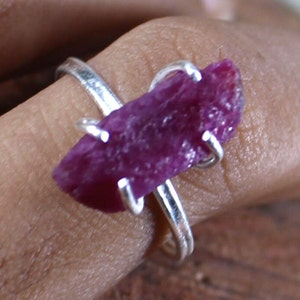 May include: A silver ring featuring a raw, oblong ruby gemstone. The ruby is a deep, rich magenta color with a textured surface. The ring's band is a simple, polished silver.