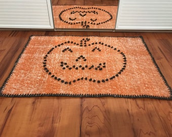 Könnte beinhalten: Orangefarbener und schwarzer Halloween-Fußabtreter mit gehäkeltem Rand und einem Kürbisgesichtsdesign. Das Kürbisgesicht besteht aus schwarzen Bommeln.