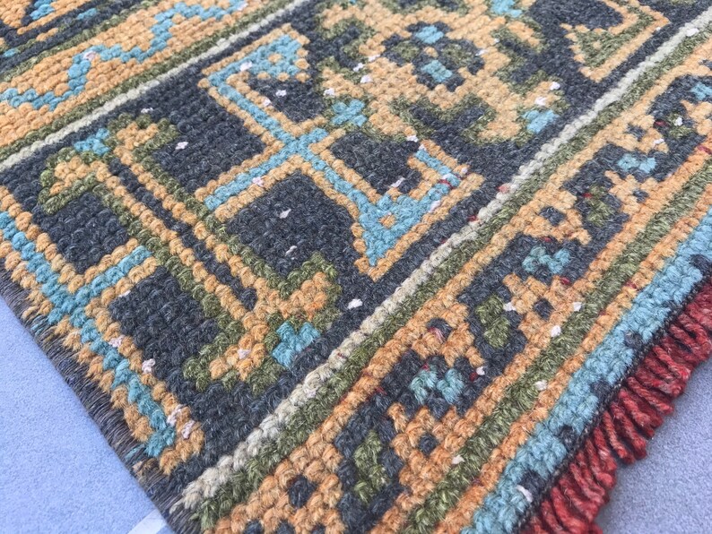 Puede incluir: Primer plano de una alfombra tejida con un patr&oacute;n geom&eacute;trico en tonos de azul, verde, marr&oacute;n y naranja. La alfombra tiene un borde de flecos.