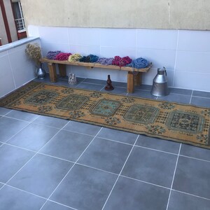 Puede incluir: Una alfombra vintage tejida a mano con un patr&oacute;n geom&eacute;trico en tonos azules, verdes y marrones. La alfombra est&aacute; colocada sobre un suelo de baldosas, con un banco de madera y contenedores de metal en el fondo.