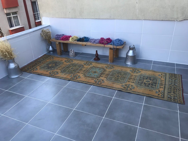 Puede incluir: Una alfombra vintage tejida a mano con un patr&oacute;n geom&eacute;trico en tonos azules, verdes y marrones. La alfombra est&aacute; colocada sobre un suelo de baldosas, con un banco de madera y contenedores de metal en el fondo.