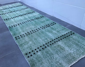 Puede incluir: Una alfombra verde y blanca con pompones negros. La alfombra tiene un aspecto desgastado y está hecha de un material suave y tejido.