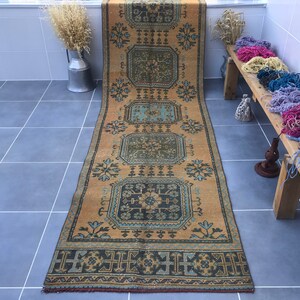 Puede incluir: Una alfombra vintage larga y estrecha con un patr&oacute;n geom&eacute;trico en tonos azules, amarillos y marrones. La alfombra tiene un borde con flecos y est&aacute; colocada sobre un suelo de baldosas.