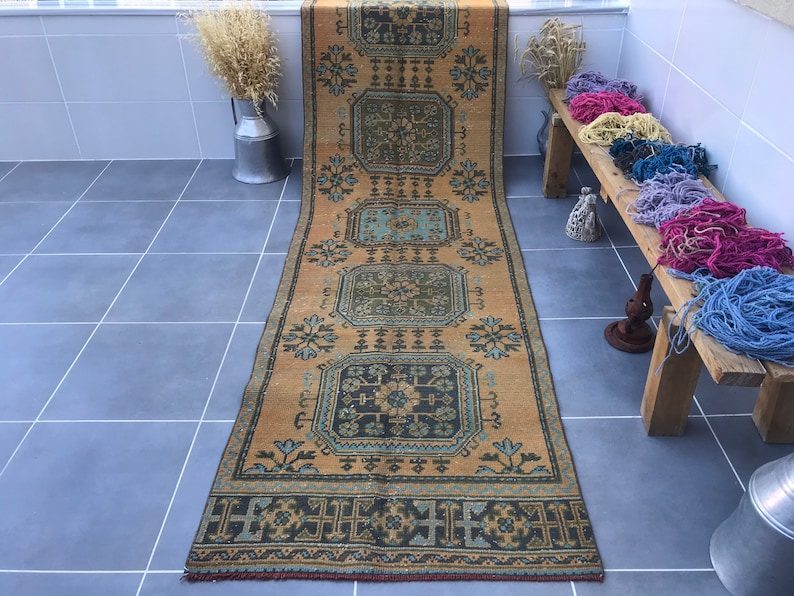 Puede incluir: Una alfombra vintage larga y estrecha con un patr&oacute;n geom&eacute;trico en tonos azules, amarillos y marrones. La alfombra tiene un borde con flecos y est&aacute; colocada sobre un suelo de baldosas.