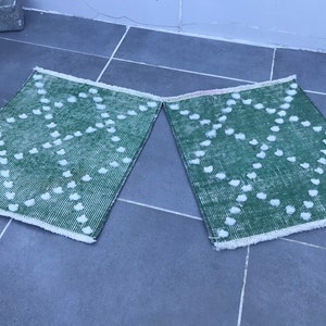 May include: Two green bath mats with white pom-poms in a geometric pattern. The mats have a white fringe border.