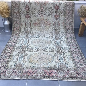 Puede incluir: Una alfombra vintage con fondo crema y intrincados patrones florales y geométricos en tonos marrones, rosas y negros. La alfombra tiene un aspecto descolorido y desgastado, lo que le da un toque único y antiguo.