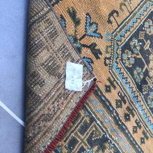 Puede incluir: Primer plano de una alfombra con un patr&oacute;n geom&eacute;trico marr&oacute;n, azul y verde. La alfombra tiene flecos rojos y una etiqueta que dice "32 x 91".