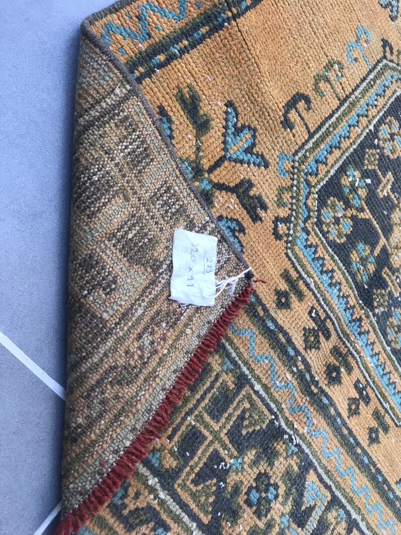 Puede incluir: Primer plano de una alfombra con un patr&oacute;n geom&eacute;trico marr&oacute;n, azul y verde. La alfombra tiene flecos rojos y una etiqueta que dice "32 x 91".