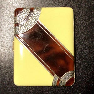 May include: A rectangular cigarette case featuring a pale yellow enamel finish with a diagonal band of dark brown tortoiseshell pattern. The corners are accented with silver-toned metal detailing that has a textured, crackled appearance.
