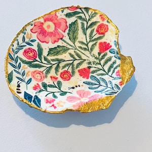 May include: A decorative seashell with a floral pattern. The shell has a white base with pink and red flowers, green leaves, and a gold-painted edge. A small bee is also visible.