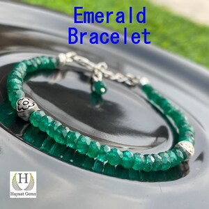 Puede incluir: Pulsera de piedras preciosas verde esmeralda con dijes de flores plateadas y un cierre de cadena plateada. La pulsera está sobre una superficie negra. "Emerald Bracelet" está escrito en azul sobre la pulsera.