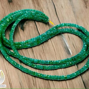 May include: A strand of teal green gemstone beads, likely aquamarine, on a wooden surface. The beads are faceted and have a slightly irregular shape. The strand is long enough to be used for a necklace or bracelet.