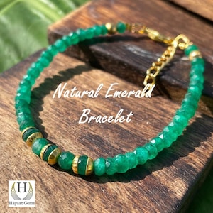 Può includere: Un bracciale con piccole perle di smeraldo verde sfaccettate alternate a distanziatori in metallo dorato liscio. Il gioiello include una catenella di estensione in metallo dorato e una chiusura. Il testo "Natural Emerald Bracelet" è visibile.
