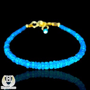 Può includere: Un braccialetto di opale blu con chiusura dorata. Il braccialetto è composto da piccole perline rotonde di opale blu chiaro. La chiusura è color oro e ha un piccolo ciondolo rotondo di opale blu chiaro. Il braccialetto è esposto su una superficie nera.