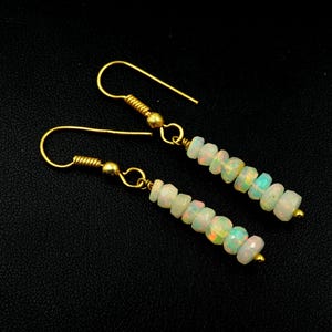 Può includere: Un paio di orecchini pendenti con piccole perline di opale sfaccettate nei toni del bianco, giallo e verde. Le perline sono infilate su un filo dorato, con un gancio dorato. Sfondo nero.
