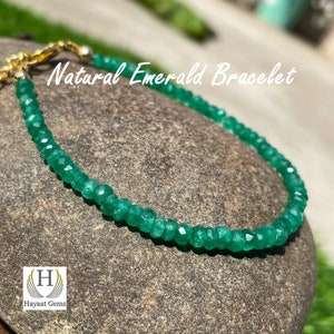 Puede incluir: Un brazalete de piedras preciosas de esmeralda verde con un cierre de oro. El brazalete está sobre una superficie de roca gris. El texto "Natural Emerald Bracelet" es visible sobre el brazalete.