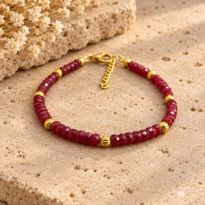 May include: A bracelet featuring faceted deep red gemstone beads alternating with small, ribbed gold-toned metal beads. The piece includes a gold-toned lobster claw clasp and an adjustable extender chain for a secure fit around the wrist.