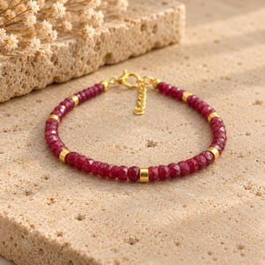 May include: A bracelet featuring small, faceted, deep red ruby beads interspersed with smooth, cylindrical gold-tone metal spacers. The piece includes a gold-tone lobster claw clasp and a short extender chain for adjustable sizing.