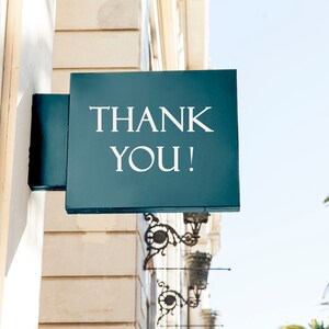 Może przedstawiać: Turkusowy znak z białym napisem "THANK YOU!", zamontowany na elewacji budynku. Znak jest kwadratowy i posiada ozdobny metalowy wspornik. W tle widoczne są palmy i czyste niebo.