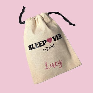 May include: A beige drawstring bag with the words "SLEEP OVER squad" printed in black and a pink glitter heart. The name "Lucy" is printed in pink glitter below the text. The bag has a black drawstring closure.