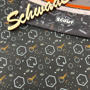 Könnte beinhalten: Schwarzer Stoff mit einem sich wiederholenden Muster aus weißen Sechsecken und orangenen Linien. Der Stoff ist bedeckt mit einer grauen Reißverschlusstasche mit dem Text "Der echte Scout" und dem Wort "Schwaaa Ba" in Holz.