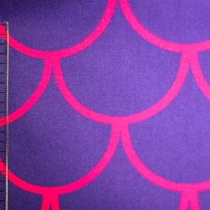 Könnte beinhalten: Lila Stoff mit einem sich wiederholenden, pinkfarbenen Muschelmuster. Ein transparenter Lineal mit Zentimetermarkierungen ist vertikal auf dem Stoff platziert. Der Stoff hat ein strukturiertes Aussehen.