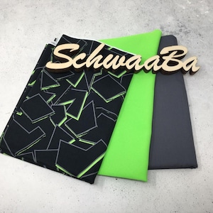 Op de afbeelding: Drie stofstalen, een zwart met een neon groen geometrisch patroon, een neon groen en een donkergrijs. De stalen zijn gestapeld met de zwarte stof bovenop. Het woord "Schwaaba" staat op een houten bord.