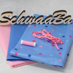 May include: Two pieces of fabric, one pink and one blue with white stars and pink polka dots, are folded together. A spool of pink thread, a pink ribbon, and a wooden sign that reads "Schwaaa Ba" are on top of the fabric.