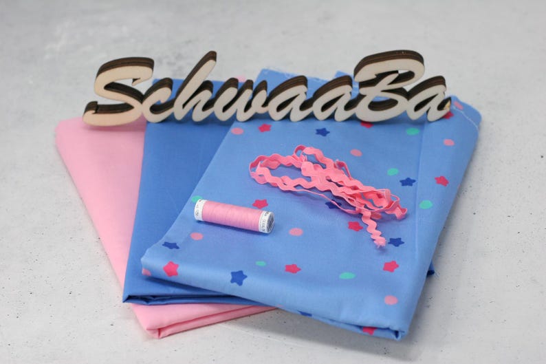 May include: Two pieces of fabric, one pink and one blue with white stars and pink polka dots, are folded together. A spool of pink thread, a pink ribbon, and a wooden sign that reads "Schwaaa Ba" are on top of the fabric.