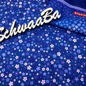Puede incluir: Una tela azul con un patrón floral blanco. La tela tiene un letrero de madera blanco con la palabra "Schwaaa Ba" en él. El letrero cubre parcialmente una bolsa de tela azul con un patrón floral blanco y el texto "Step by Step" en ella.
