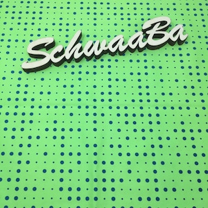 Könnte beinhalten: Ein Holzschild mit der Aufschrift "SchwaaBa" in einer stilisierten Schriftart, mit einer weißen Deckschicht und einem dunkelbraunen Schatten. Das Schild befindet sich auf einem hellgrünen Stoff mit einem Muster aus blauen Punkten.