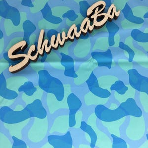 Op de afbeelding: Blauw en turquoise abstract camouflage stof met de tekst "Schwaaa Ba" in wit op een houten bord.