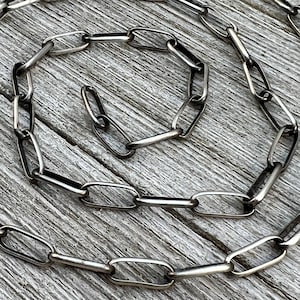Puede incluir: Un collar de cadena plateada con eslabones ovalados alargados. La cadena se muestra sobre una superficie de madera envejecida, creando un contraste entre el metal brillante y el fondo rústico. Los eslabones están conectados en un patrón repetitivo.