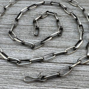 Puede incluir: Un collar de cadena plateada con eslabones ovalados y un cierre de gancho. La cadena se muestra sobre una superficie de madera gris y desgastada. El collar es una joya.