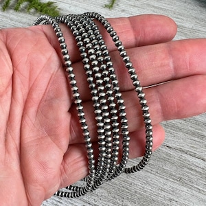 May include: A silver beaded necklace with multiple strands. The necklace is composed of small, round, silver beads strung together. The beads have a slightly oxidized finish, giving them a vintage look. The necklace is held in a hand.