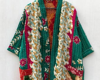May include: A green, floral-patterned kimono-style jacket with short sleeves. The jacket features a patchwork design with white, yellow, and pink floral patterns. It has two front pockets and is hanging on a wooden hanger.