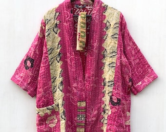 May include: A vibrant pink kimono-style robe with a patchwork design. The robe features floral and geometric patterns in cream, black, and green. It has short sleeves and two pockets. The robe is hanging on a wooden hanger.