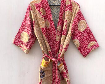 May include: A patterned robe in shades of red and beige hangs from a wooden hanger. The robe features a repeating pattern of small red shapes on a beige background, with a contrasting red trim and belt. The robe is made of a soft, flowing fabric.