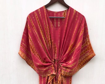 May include: A flowing, tie-dye style dress in shades of red and orange hangs from a wooden hanger. The dress features a V-neckline and a cinched waist with a drawstring. The fabric has a vertical striped pattern.