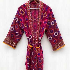 Op de afbeelding: Een bordeauxrode geborduurde kimono met een geometrisch patroon. De kimono heeft ingewikkelde stiksels in roze, blauwe en gouden tinten, met spiegelaccenten. Het heeft lange mouwen, een ceintuur en oranje afwerking. De kimono hangt aan een houten kleerhanger.