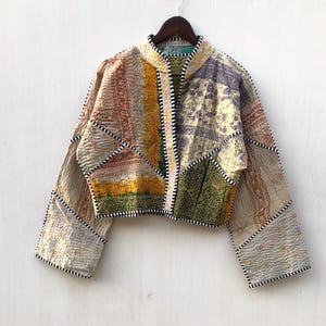 May include: A cropped jacket constructed from quilted fabric panels in various patterns and colours including yellow, green, and purple. The garment features a mandarin collar and long sleeves, with all edges finished in a black and white striped trim.