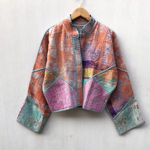 May include: A cropped jacket featuring a patchwork design with orange, pink, purple, and teal colors. The garment has long sleeves, a high collar, and visible white stitching throughout the fabric, hanging on a wooden hanger.
