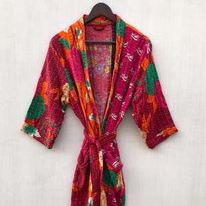 May include: A vibrant, quilted robe with a floral pattern in shades of red, orange, and green. The robe has a tie closure and short sleeves. The garment is hanging on a wooden hanger against a white background.