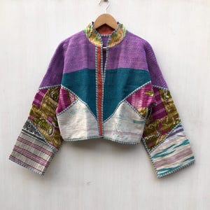 Könnte beinhalten: Eine kurz geschnittene Jacke mit Patchwork-Design aus verschiedenen Stoffmustern in Violett, Petrol, Weiß und Olivgrün. Das Kleidungsstück hat lange Ärmel, einen Stehkragen und sichtbare Nähte über die gesamte gesteppte Konstruktion hinweg.