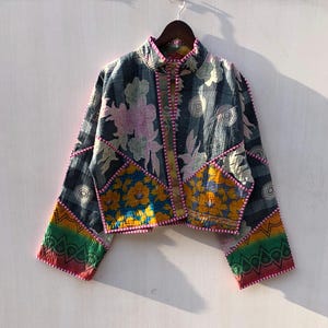 Puede incluir: Chaqueta patchwork con cuello alto y mangas largas. La chaqueta presenta una mezcla de estampados florales y geom&eacute;tricos en tonos azules, amarillos, rosas y verdes, con ribete rosa. Cuelga de una percha de madera.