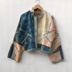 May include: A cropped jacket featuring a patchwork design of teal and beige fabric panels. The garment has long sleeves, a mandarin collar, and visible white contrast stitching along the seams and edges, hanging on a wooden hanger.