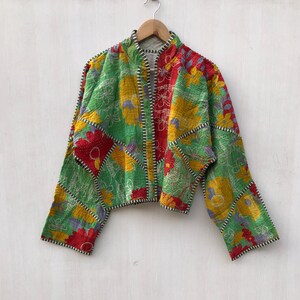 May include: A cropped jacket featuring a patchwork design of floral patterns in vibrant shades of green, yellow, and red. The garment has long sleeves, a stand-up collar, and visible white stitching along the seams, hanging on a wooden hanger.